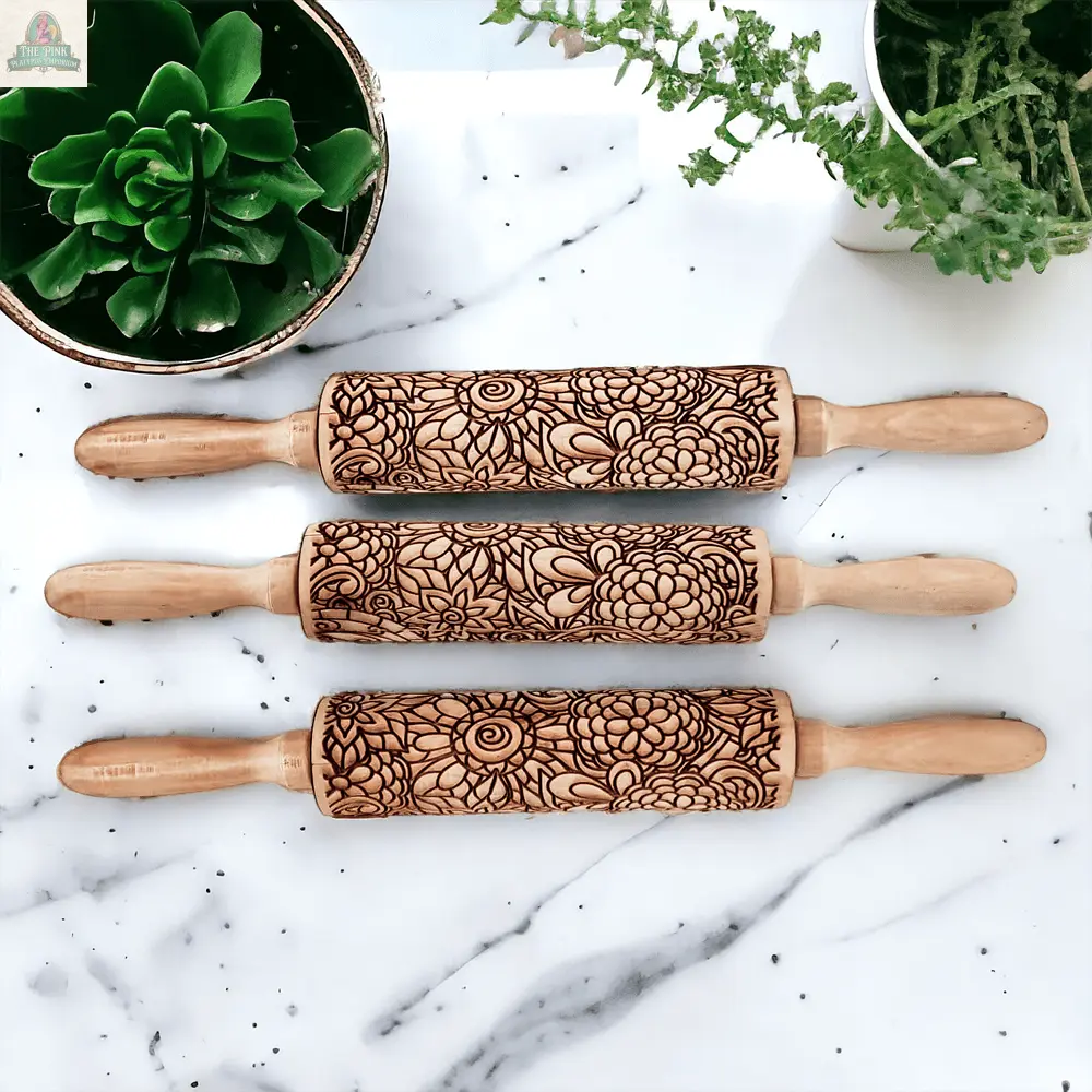 Three Rolling Pins - Box Style with detailed floral carvings rest on a white marble surface, creating a charming baking gift. Two green potted plants in the corners accentuate the artisan rolling pins beautifully.