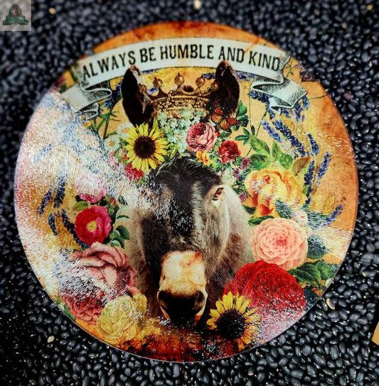 The large 12" round glass cutting board features a crowned donkey among flowers and greenery with “Always Be Humble and Kind” on top, resting on black beans for a fun charcuterie or trivet display.