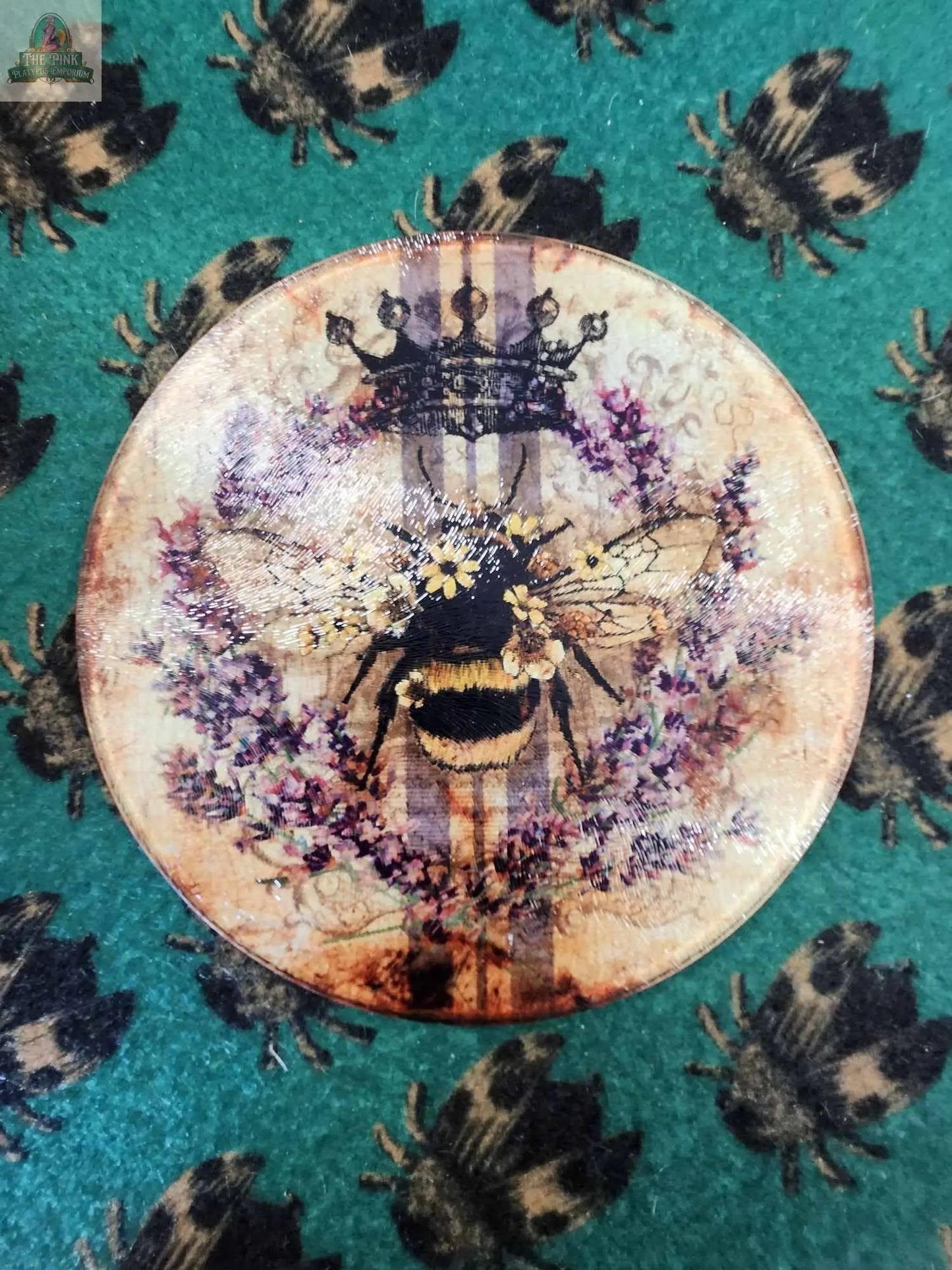 A 12" round glass cutting board with a vintage bee and lavender design, featuring a crown and purple flowers, rests on green fabric with a repeating bee pattern.