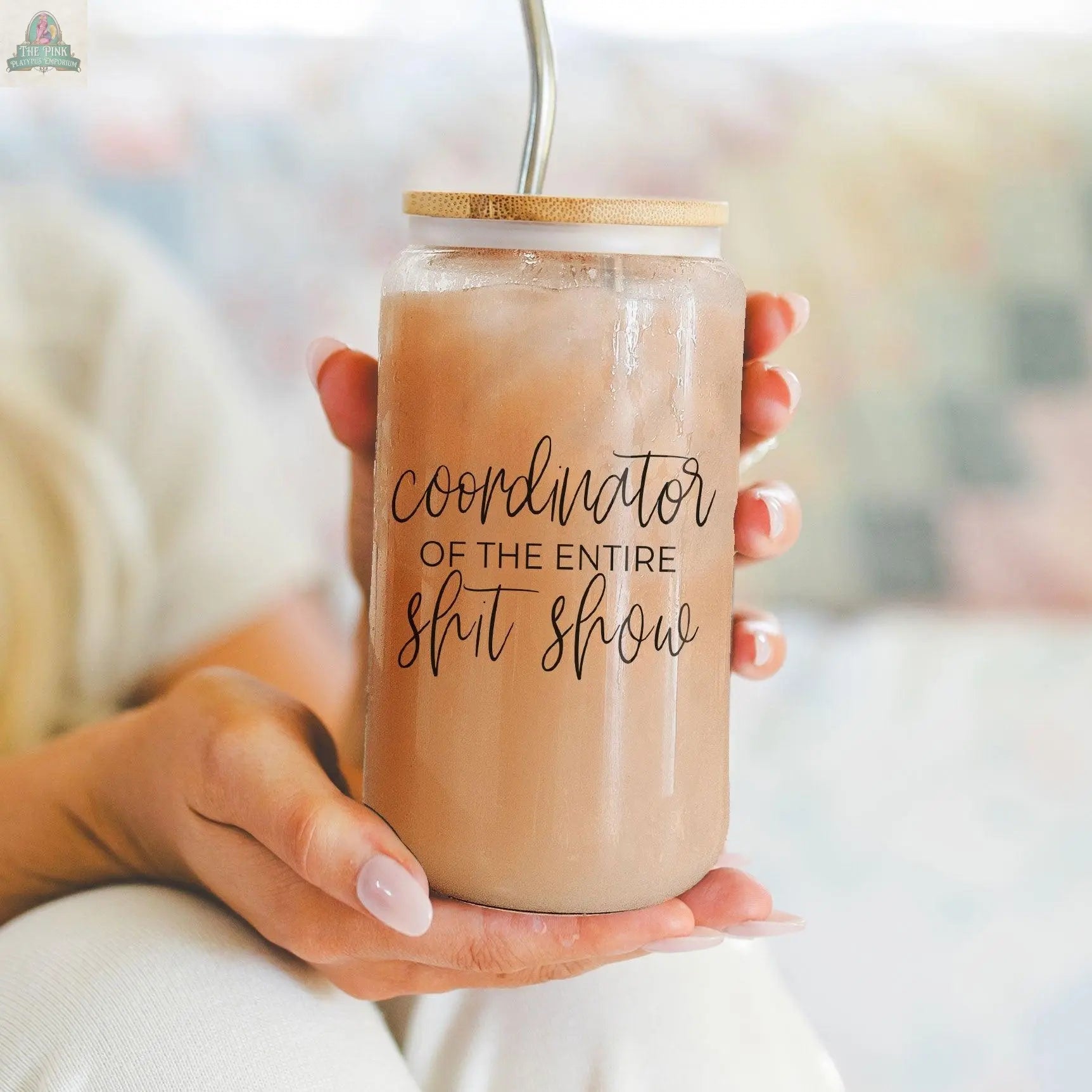 A person holds the S**t Show Coordinator 17oz glass jar with iced coffee and a stainless steel straw. The text on the jar reads “Coordinator of the entire shit show,” and the softly blurred background adds focus to the cup.