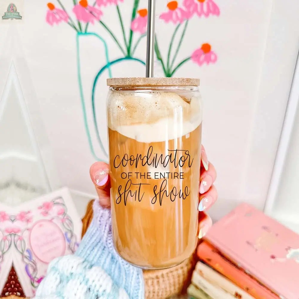 A hand holds the S**t Show Coordinator 17oz glass cup filled with iced coffee and a stainless steel straw, set against a backdrop of books, a cozy knitted blanket, and a flower drawing in a vase.