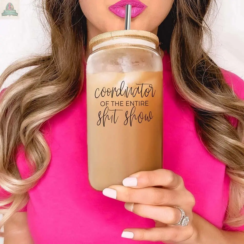 A woman with wavy hair and manicured nails holds the S**t Show Coordinator 17oz glass cup filled with iced coffee and a stainless steel straw. She wears a diamond ring and a bright pink top.