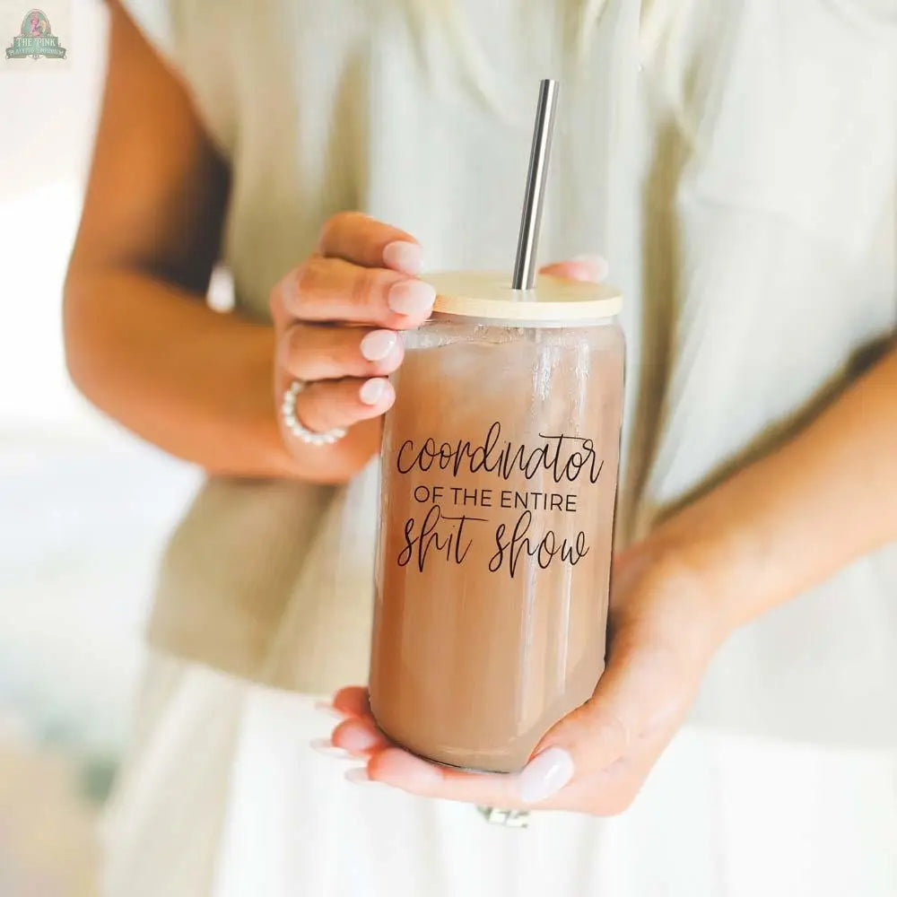 A woman in a light shirt holds the S**t Show Coordinator 17oz glass cup, filled with iced coffee and topped with a bamboo lid and stainless steel straw. "Coordinator of the entire sh*t show" is printed in decorative script on the glass.