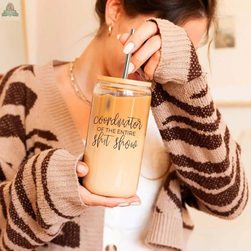 A woman in a brown and beige sweater holds iced coffee in the S**t Show Coordinator 17oz glass with a stainless steel straw. Her face is partly covered by her sleeve.