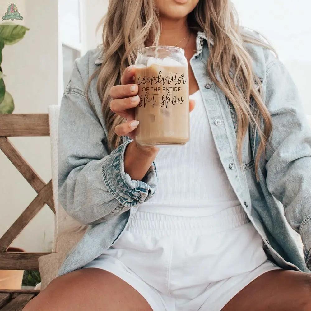 A woman in a denim shirt and white shorts sits on a bench, holding the S**t Show Coordinator 17oz glass cup with bamboo lid and stainless steel straw.