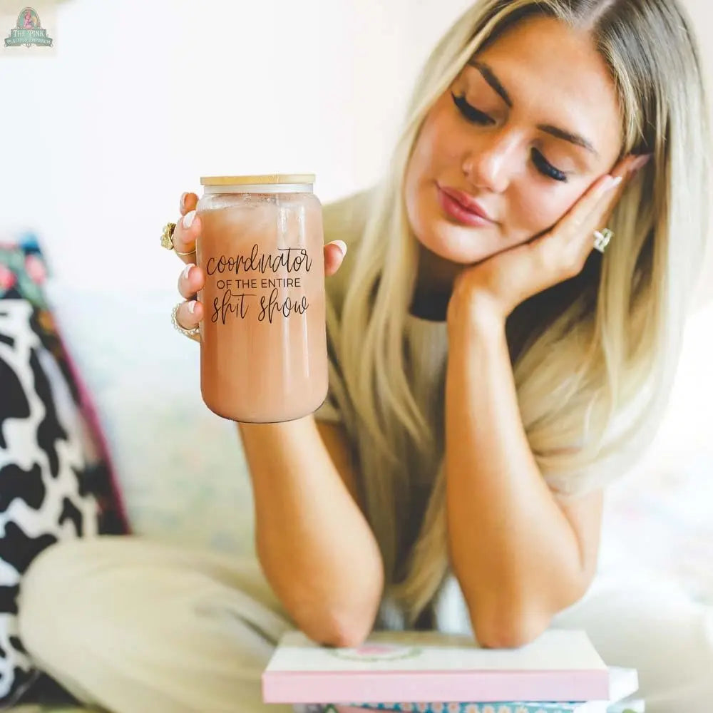A woman with long blonde hair sits with her head resting on her hand, holding the S**t Show Coordinator 17oz mason jar with a stainless steel straw, looking thoughtful as a book rests on her lap.