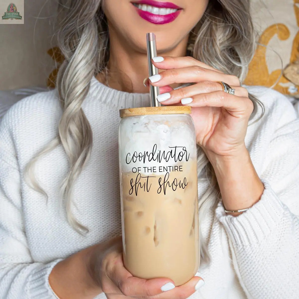 A woman with long, wavy gray hair and bright pink lipstick holds an iced latte in the S**t Show Coordinator 17oz glass with a stainless steel straw. The cup reads “Coordinator of the entire shit show” in black script.