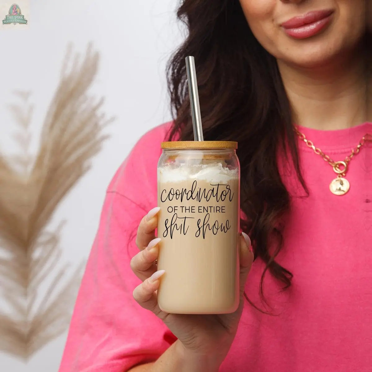 A woman in a pink shirt holds the S**t Show Coordinator 17oz glass filled with iced coffee, whipped cream, and a stainless steel straw; the glass features "Coordinator of the entire sh*t show" in black lettering.