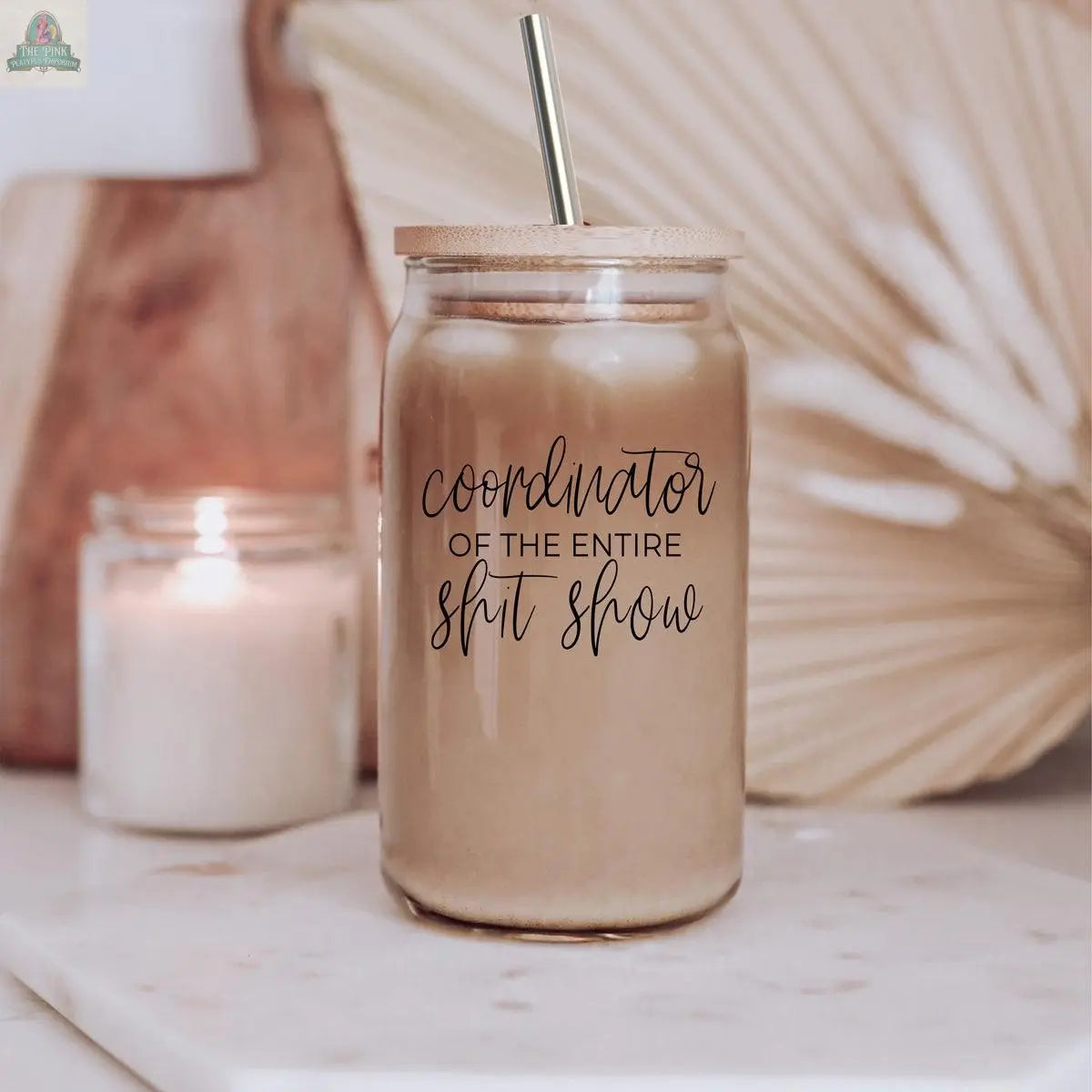 A S**t Show Coordinator 17oz glass cup with iced coffee and a stainless steel straw sits on the counter. The jar reads "Coordinator of the entire shit show," while a lit candle and decor blur in the background.
