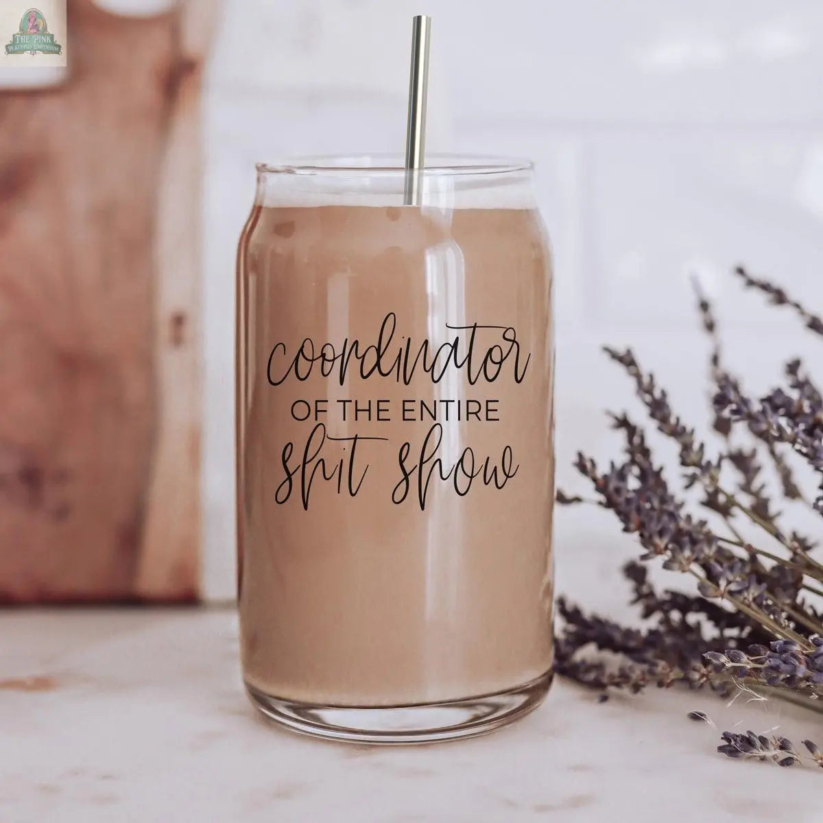 A 17oz S**t Show Coordinator cup with a bamboo lid and stainless steel straw holds iced coffee on the counter; script-style label visible, with lavender sprigs and a wooden board in the background.
