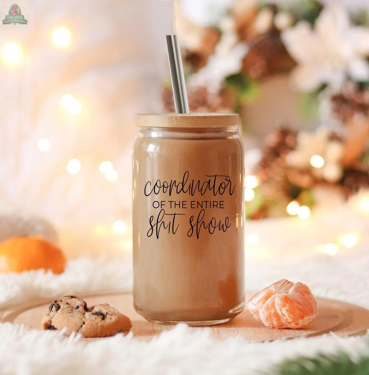 A S**t Show Coordinator 17oz glass jar with a stainless steel straw and bamboo lid sits on a tray with chocolate chip cookies and a peeled orange, soft lights, and festive decor in the background.