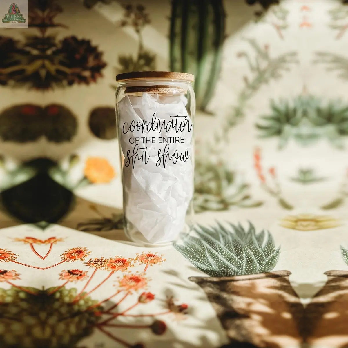 The S**t Show Coordinator 17oz glass jar with a cork lid, filled with crumpled white paper, sits on a botanical table beside glass cups. The jar’s label reads: Coordinator of the entire shit show.