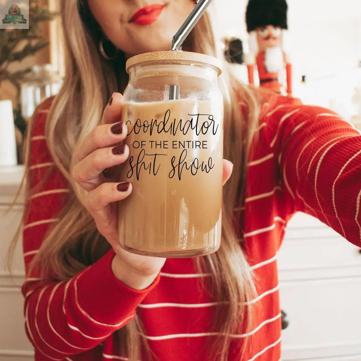 A woman in a red and white striped sweater holds a S**t Show Coordinator 17oz glass with iced coffee and a stainless steel straw. Her lips are painted red.