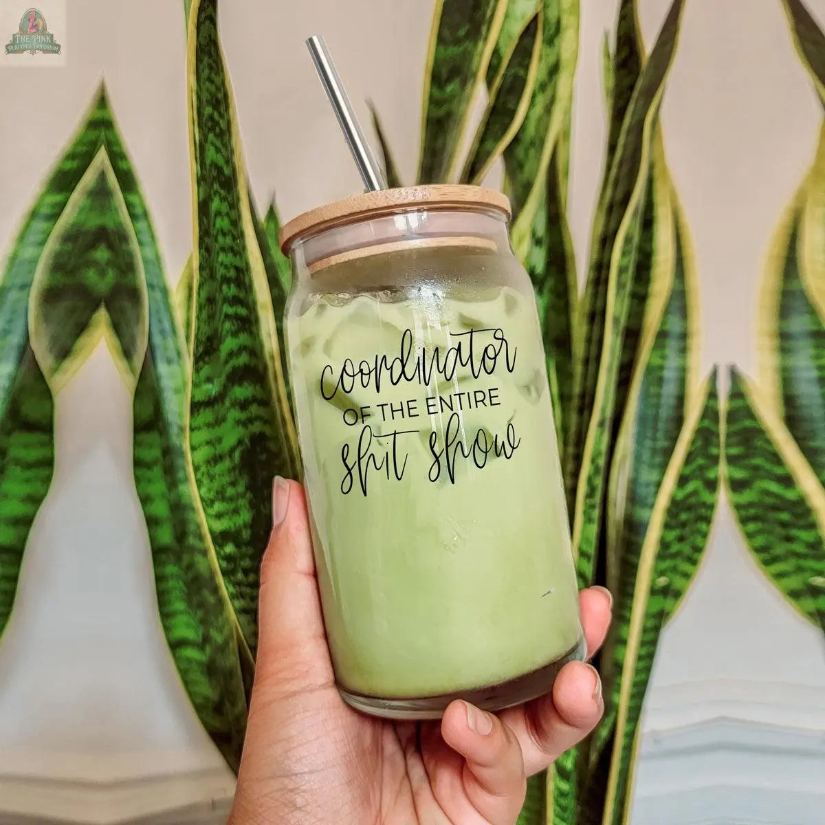 A hand holds the S**t Show Coordinator 17oz bamboo lid cup filled with a green iced drink and a stainless steel straw, with tall green snake plants in the background.