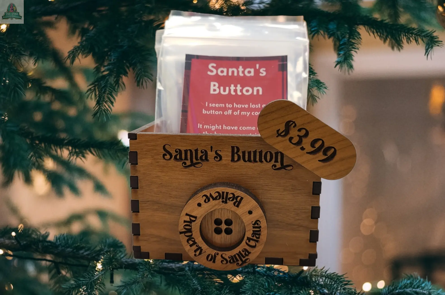 A wooden display box labeled "Santa's Button" sits among Christmas branches, showing a $3.99 price and containing the Santa's Button package—an ideal, charming Christmas gift.