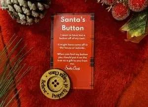 The Santa's Button, a round tan button labeled “Believe, Magic, Proven,” is paired with a red “Santa’s Button” card featuring a poem. The festive scene includes pinecones, greenery, red berries, and an ornament on soft red fabric.