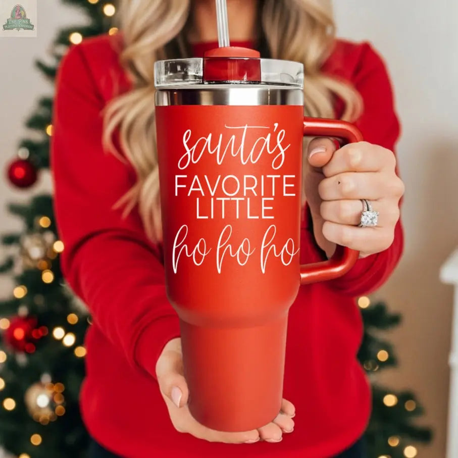 A woman in a red sweater holds a Santa's Ho 40oz mug with "Santa’s Favorite Little Ho Ho Ho" printed on it, standing in front of a decorated Christmas tree.