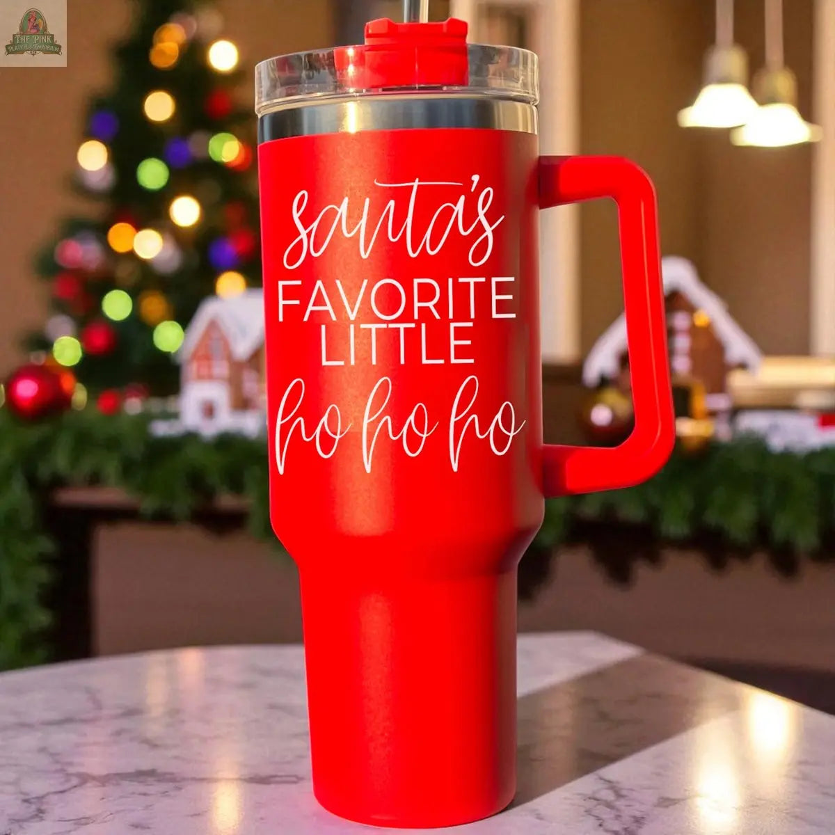 A 40oz red "Santa's Ho 40oz" mug with “Santas favorite little ho ho ho” in white script rests on a marble surface, while a decorated Christmas tree and gingerbread houses create a festive backdrop.
