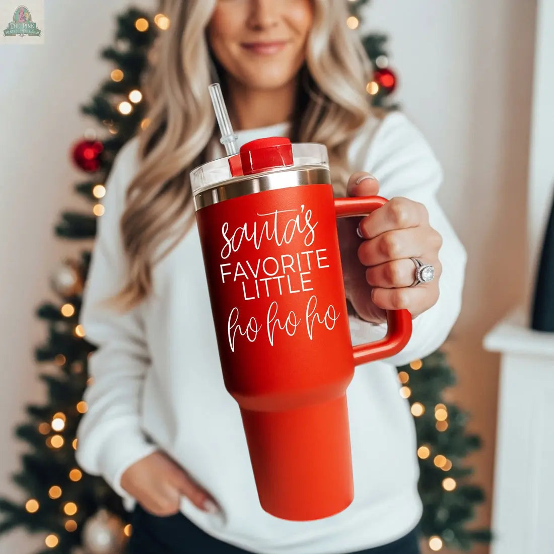 A woman in a white sweater stands before a decorated Christmas tree, holding the Santa's Ho 40oz red holiday mug featuring a handle and the phrase "Santas Favorite Little Ho Ho Ho.
