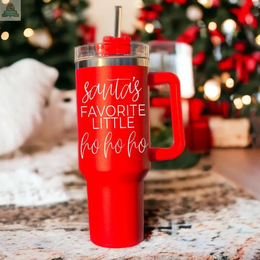 The Santa's Ho 40oz red Christmas tumbler with a handle and straw rests on a rug. “Santas Favorite Little Ho Ho Ho” is printed in white, while a blurred Christmas tree and gifts create a festive background.