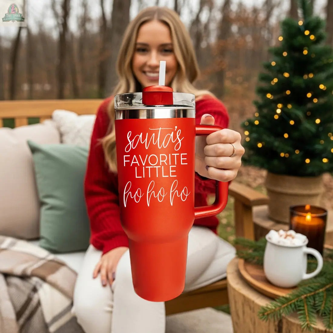 A smiling woman in a red sweater sits on an outdoor bench, holding the Santa's Ho 40oz mug. Beside her are a small lit Christmas tree and a mug of hot chocolate.