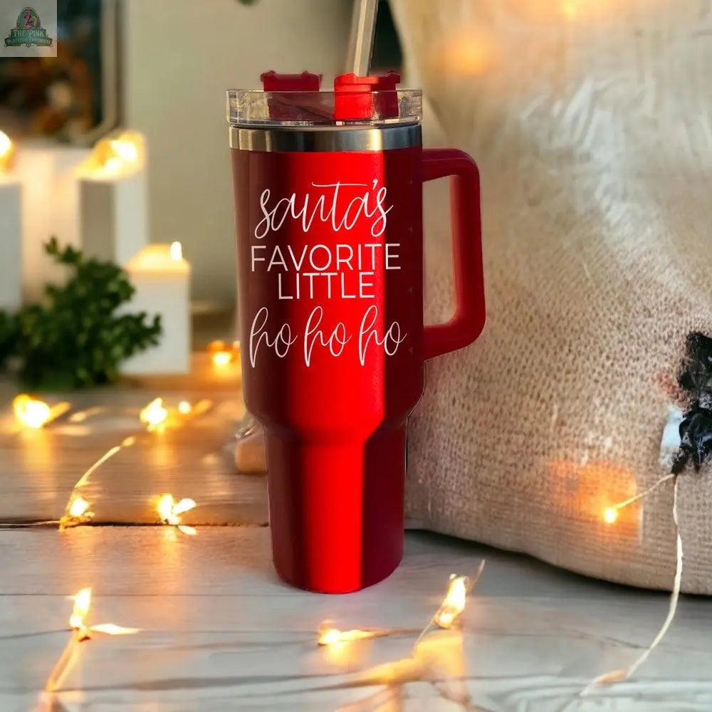 The Santa's Ho 40oz red insulated mug with handle and straw sits on a wooden surface with string lights. It features “Santas Favorite Little Ho Ho Ho” in white script, perfect for holiday cheer.
