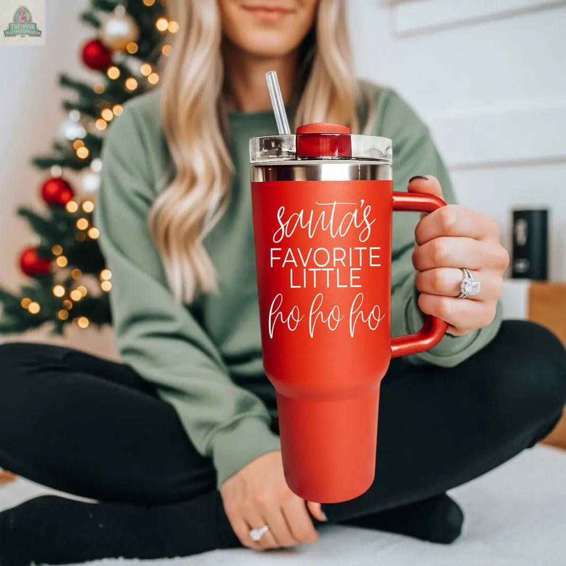 A woman with long blonde hair in a green sweater and black pants sits cross-legged by a Christmas tree, holding the Santa's Ho 40oz red tumbler labeled "Santa's favorite little ho ho ho.