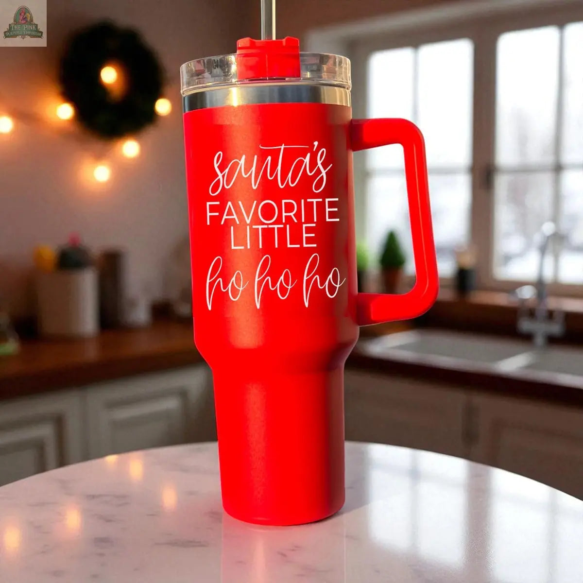A Santa's Ho 40oz red insulated mug with handle and straw sits on a kitchen counter, reading "Santa’s favorite little ho ho ho" in white script. A festive kitchen with warm lighting and a holiday wreath completes the holiday scene.