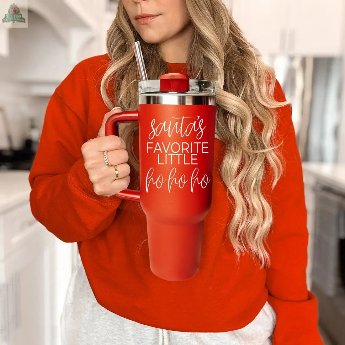A woman in a red sweater stands in a kitchen, holding the Santa's Ho 40oz insulated mug with straw, which features "Santas Favorite Little Ho Ho Ho" in white script—a perfect festive Christmas tumbler.