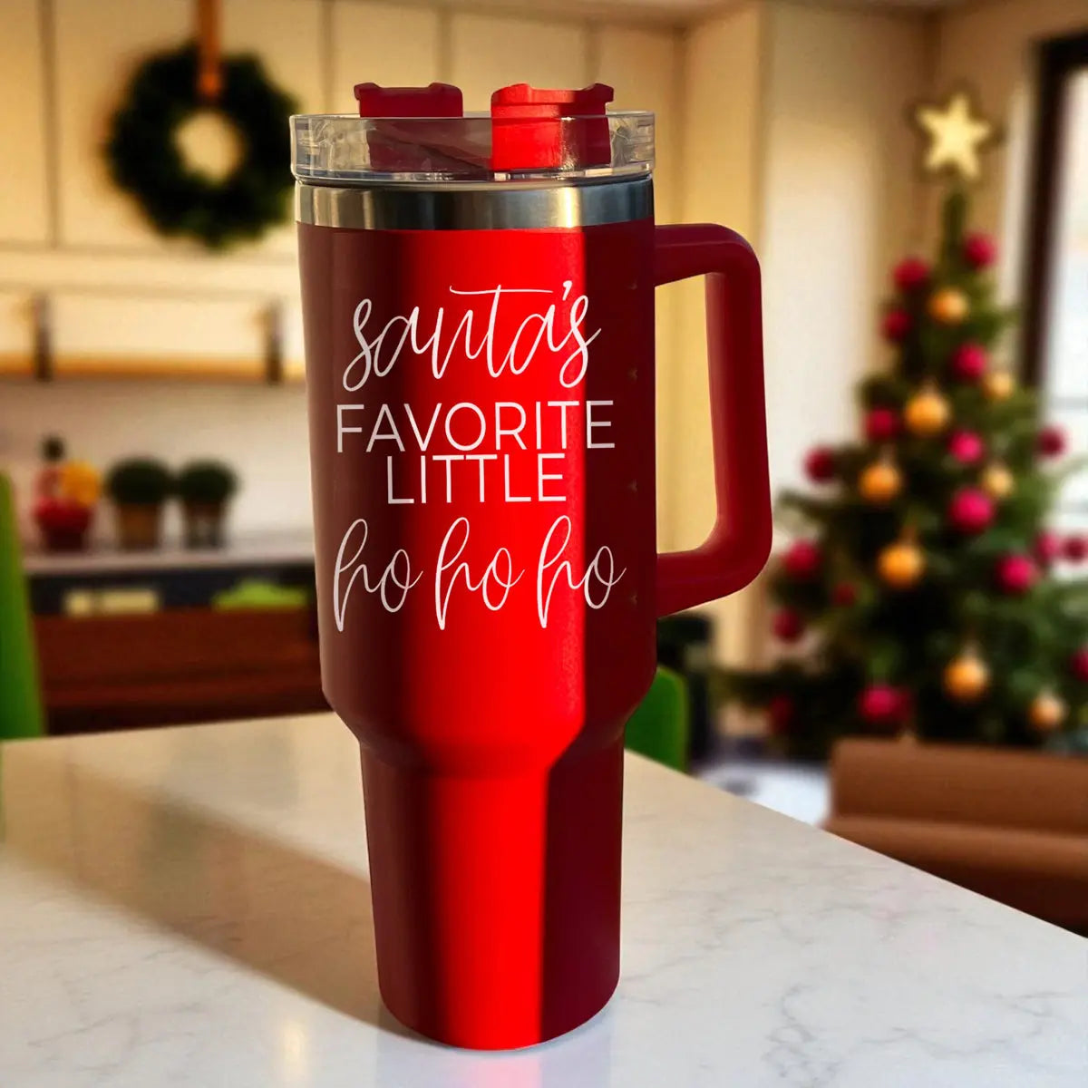 The Santa's Ho 40oz holiday mug with lid rests on a kitchen counter, featuring “Santas favorite little ho ho ho” in white script. A decorated Christmas tree and kitchen enhance the festive charm of this Christmas tumbler.