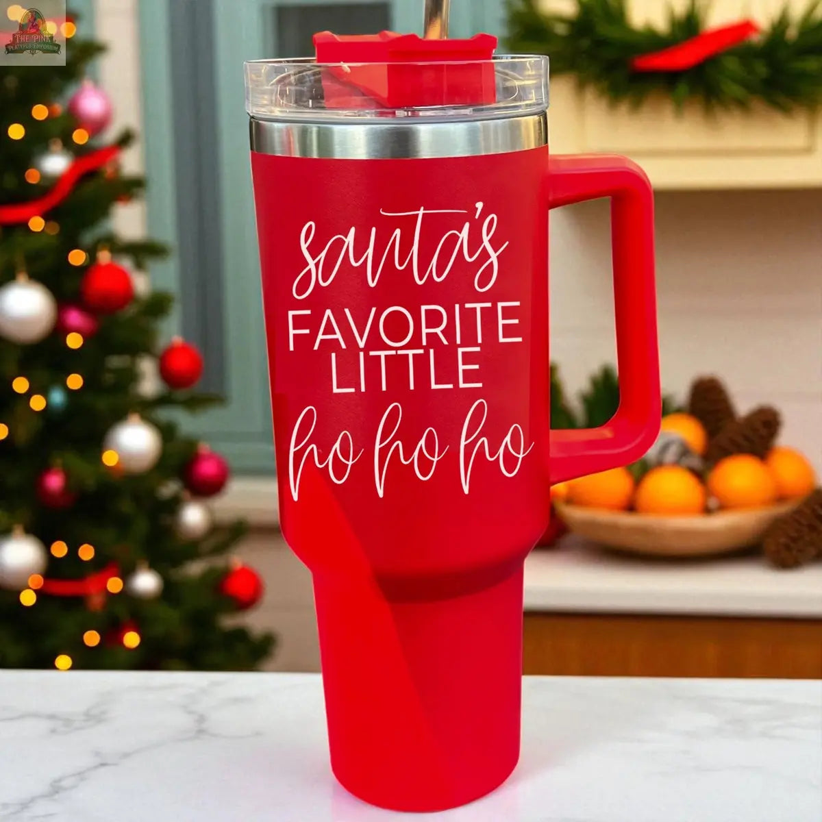 The Santa's Ho 40oz red holiday mug, with a lid and handle, shows white “Santa’s FAVORITE LITTLE ho ho ho.” text. It’s pictured on a kitchen counter with festive decor and a Christmas tree in the background.