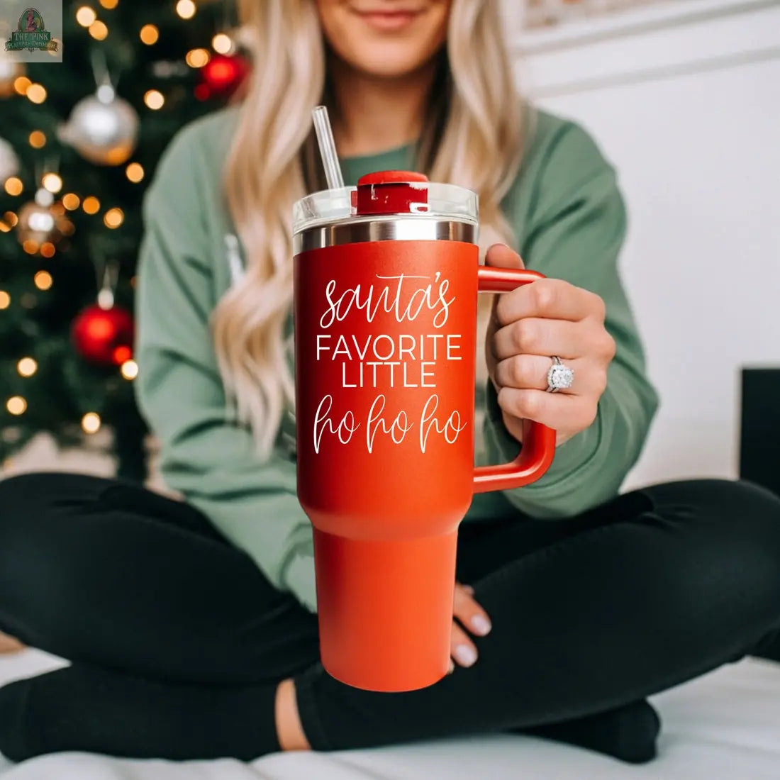A woman in a green top with an engagement ring sits cross-legged by a Christmas tree, holding the Santa's Ho 40oz holiday mug with straw that reads "Santa’s favorite little ho ho ho.