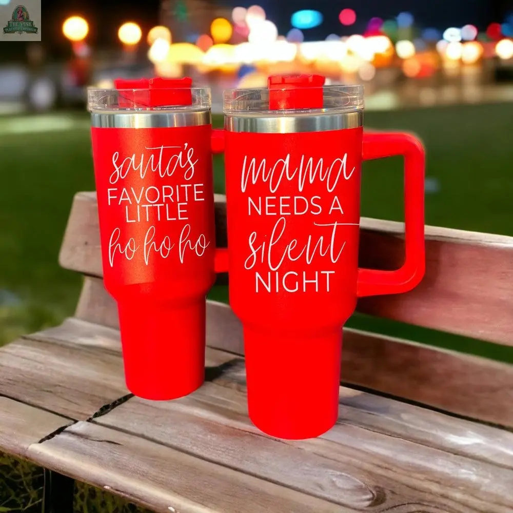 Two red Santa's Ho 40oz Christmas tumblers sit on a wooden bench—one says "santas favorite little ho ho ho," the other reads "mama needs a silent night." Colorful blurred lights in the background set a festive holiday mood.