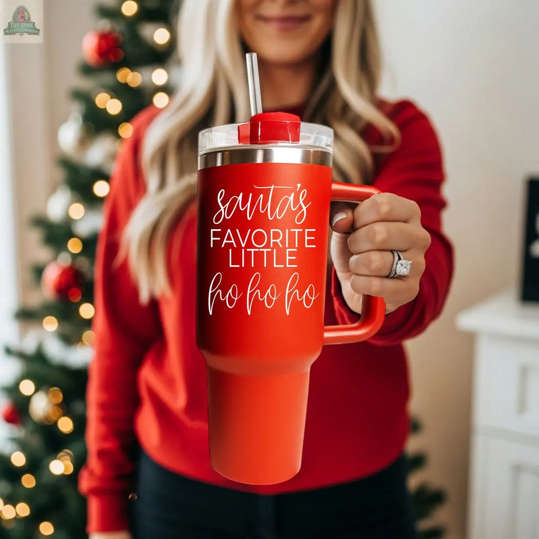 A woman in a red sweater stands by a decorated Christmas tree, holding the Santa's Ho 40oz holiday mug with a straw that reads “Santa’s favorite little ho ho ho.”.