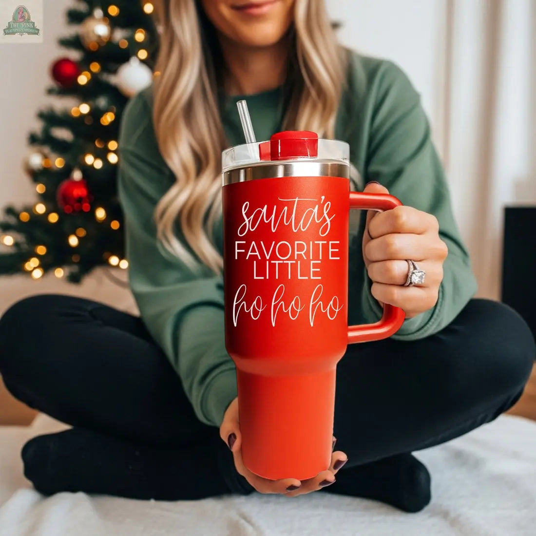 A woman in a green sweater sits by a decorated Christmas tree, holding a large red 40oz "Santa's Ho" holiday mug with a straw and the words "Santa’s Favorite Little Ho Ho Ho" printed in white.