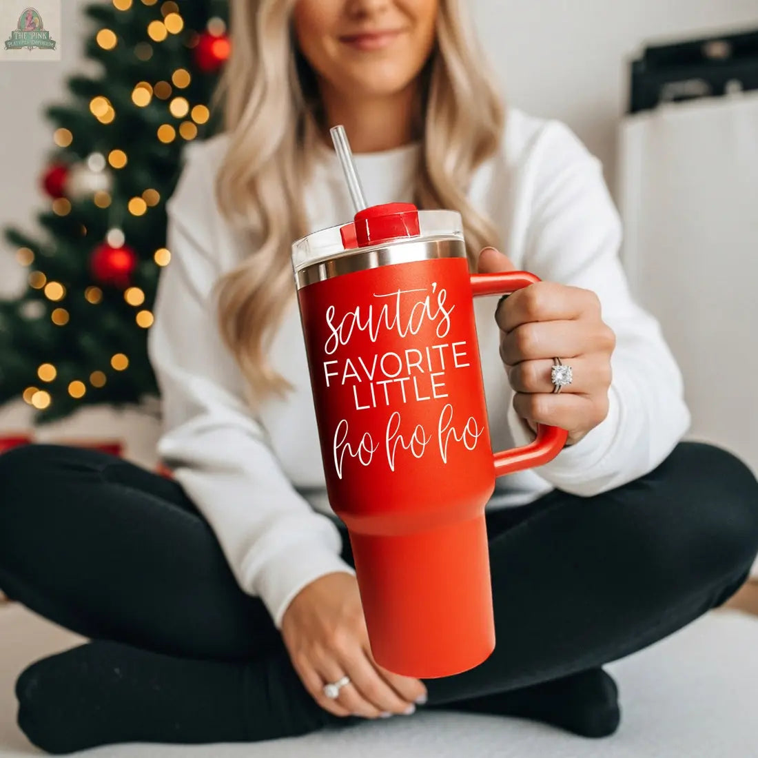 A woman with long blonde hair holds the Santa's Ho 40oz red holiday mug with "Santa’s favorite little ho ho ho" in white script, sitting cross-legged by a decorated Christmas tree in a white sweater and black pants.