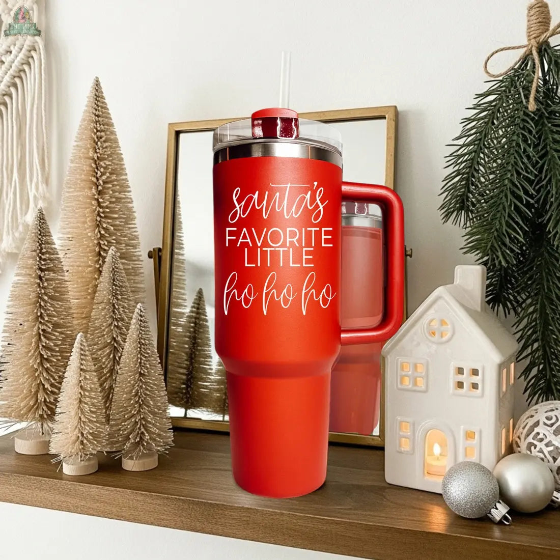 The Santa's Ho 40oz red Christmas tumbler with a clear lid and straw says “Santa’s favorite little ho ho ho.” It rests on a wooden shelf next to mini Christmas trees, a lit ceramic house, pine branch, and reflects in a mirror.