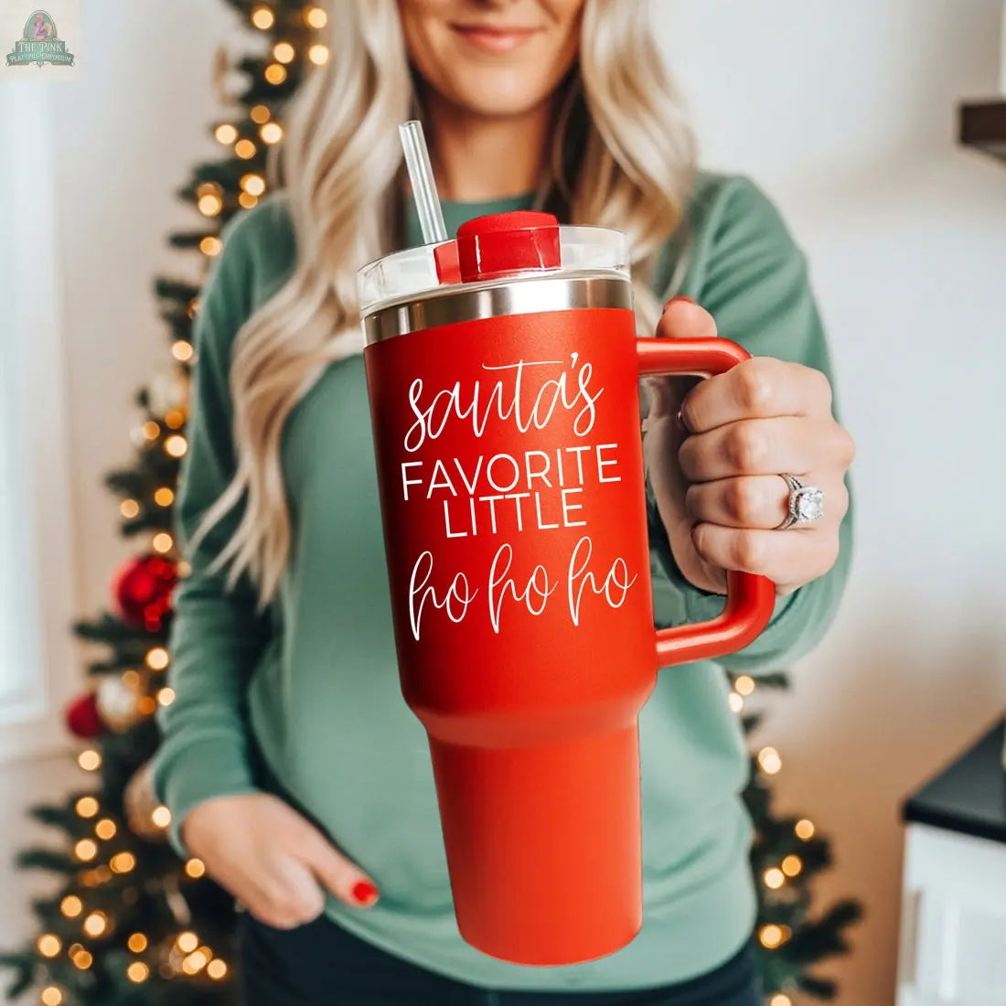 A woman in a green top holds the Santa's Ho 40oz holiday mug with handle and straw, featuring "Santa's favorite little ho ho ho." A blurred Christmas tree decorates the background.