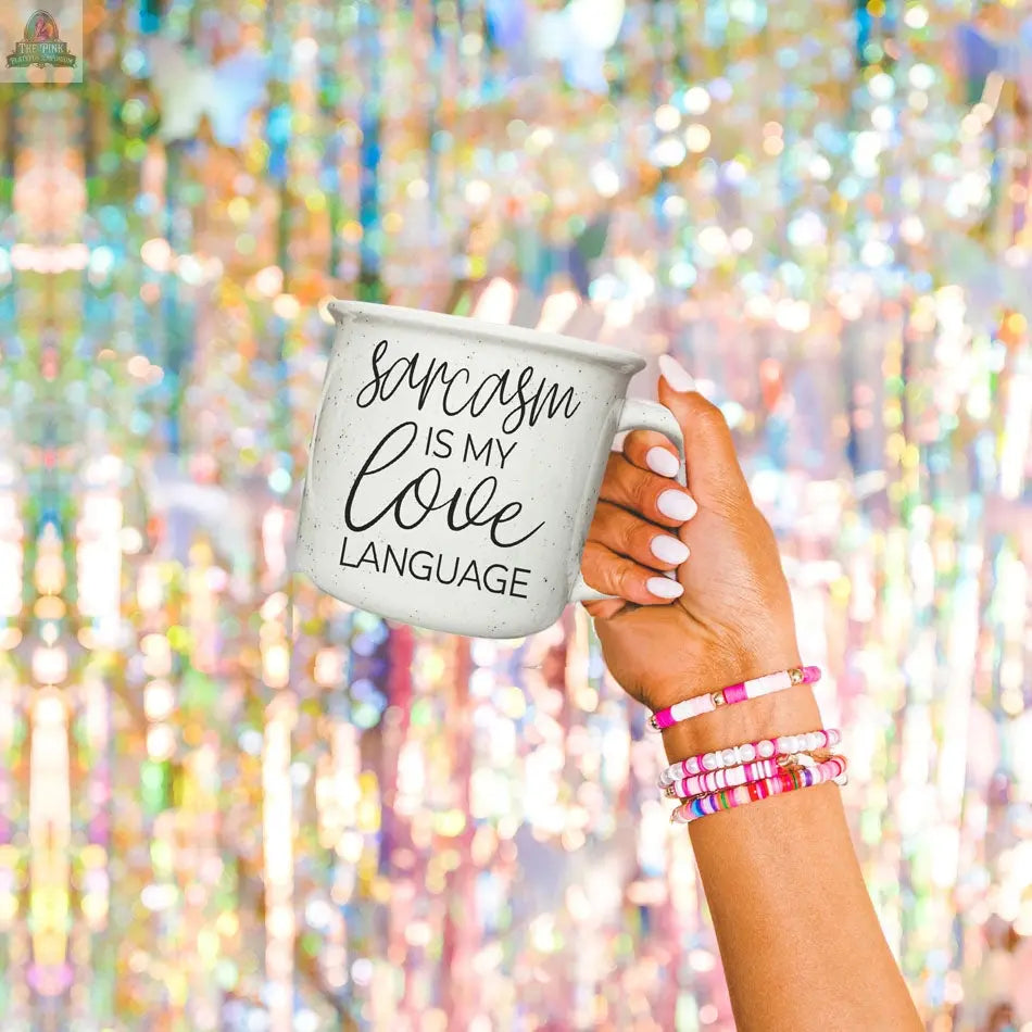 A hand with colorful beaded bracelets holds the Sarcasm Love 14.5oz ceramic mug with “Sarcasm is my love language” in black script, set against a sparkling, multicolored, festive background.