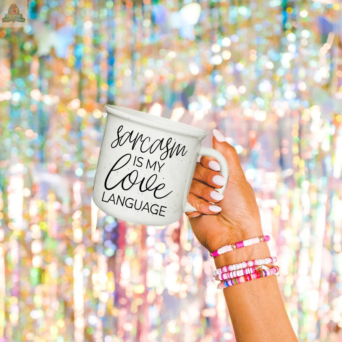 A hand with colorful beaded bracelets holds the Sarcasm Love 14.5oz ceramic mug, which features “Sarcasm is my love language” against a sparkling, iridescent background.