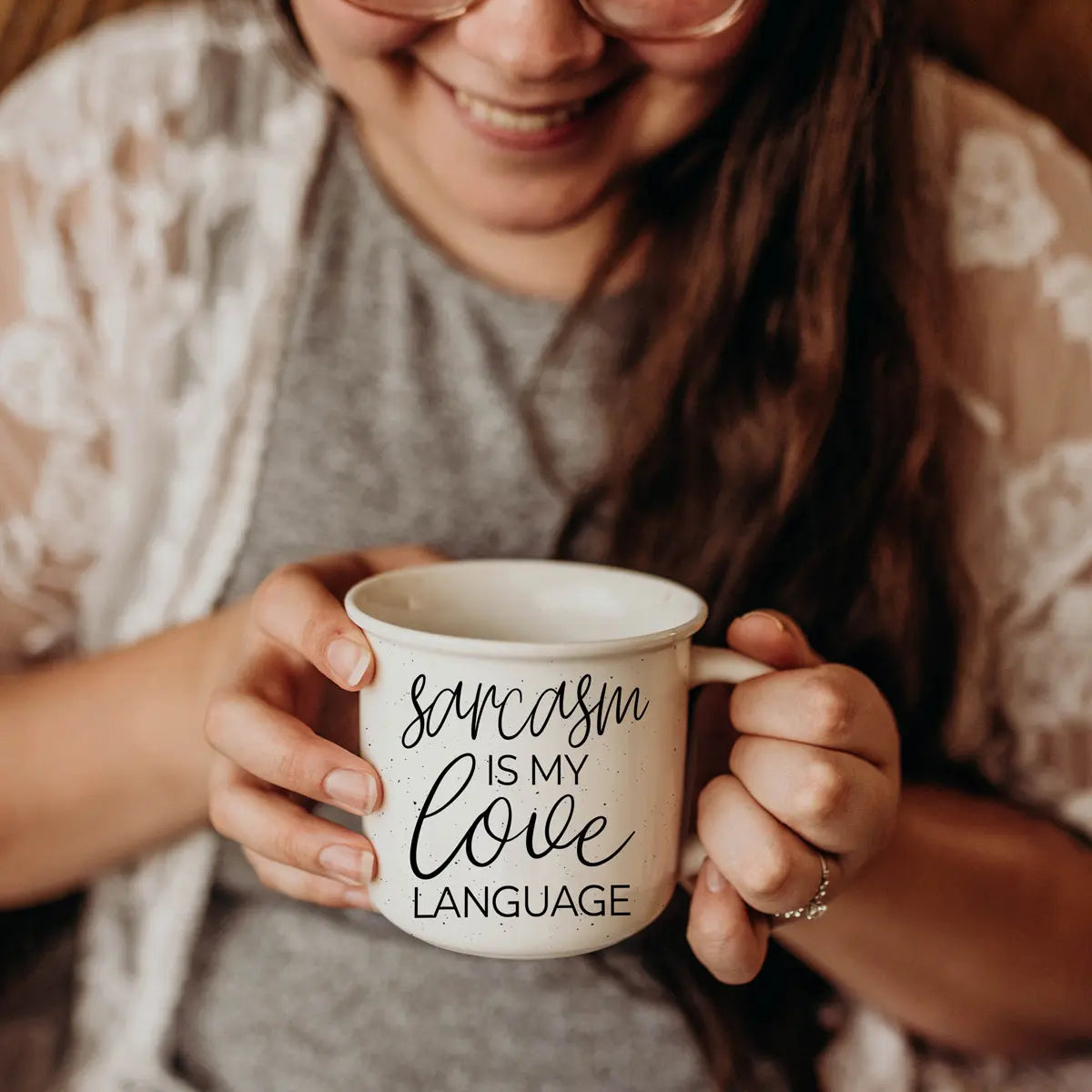 A smiling person holds the Sarcasm Love 14.5oz mug that says "Sarcasm is my love language," showing off this funny coffee mug—ideal for anyone fluent in sarcasm. They wear glasses, a gray shirt, and a light lace cardigan.