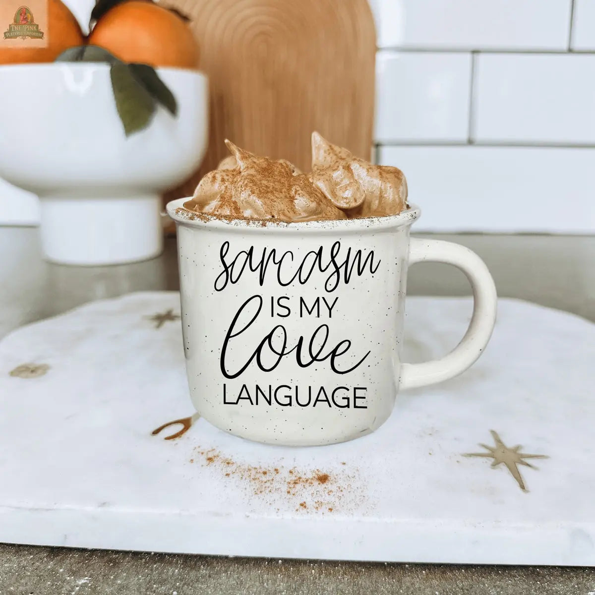A 14.5oz Sarcasm Love mug filled with whipped coffee rests on a marble board. The funny ceramic mug reads “Sarcasm is my love language,” with a bowl of oranges and a white subway tile backsplash in the background.