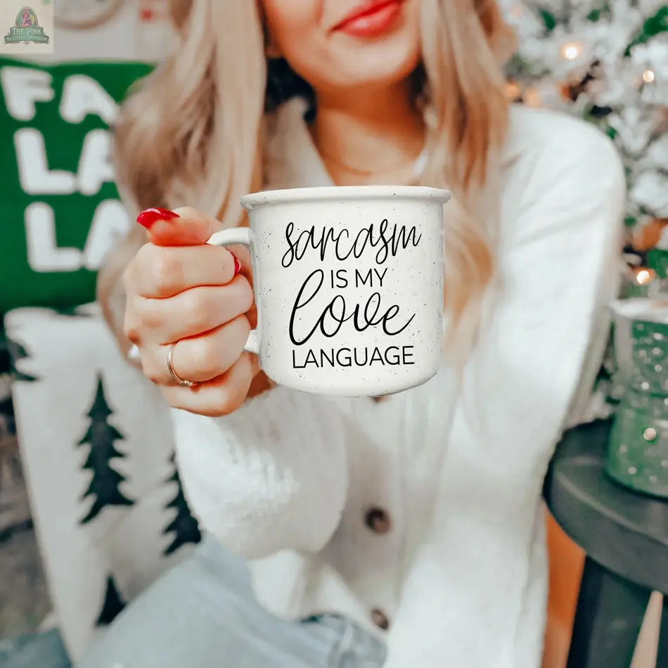 A woman with long blonde hair holds the Sarcasm Love 14.5oz mug toward the camera. The white ceramic mug shows the black text, “Sarcasm is my love language,” with a Christmas tree and holiday decor in the festive background.