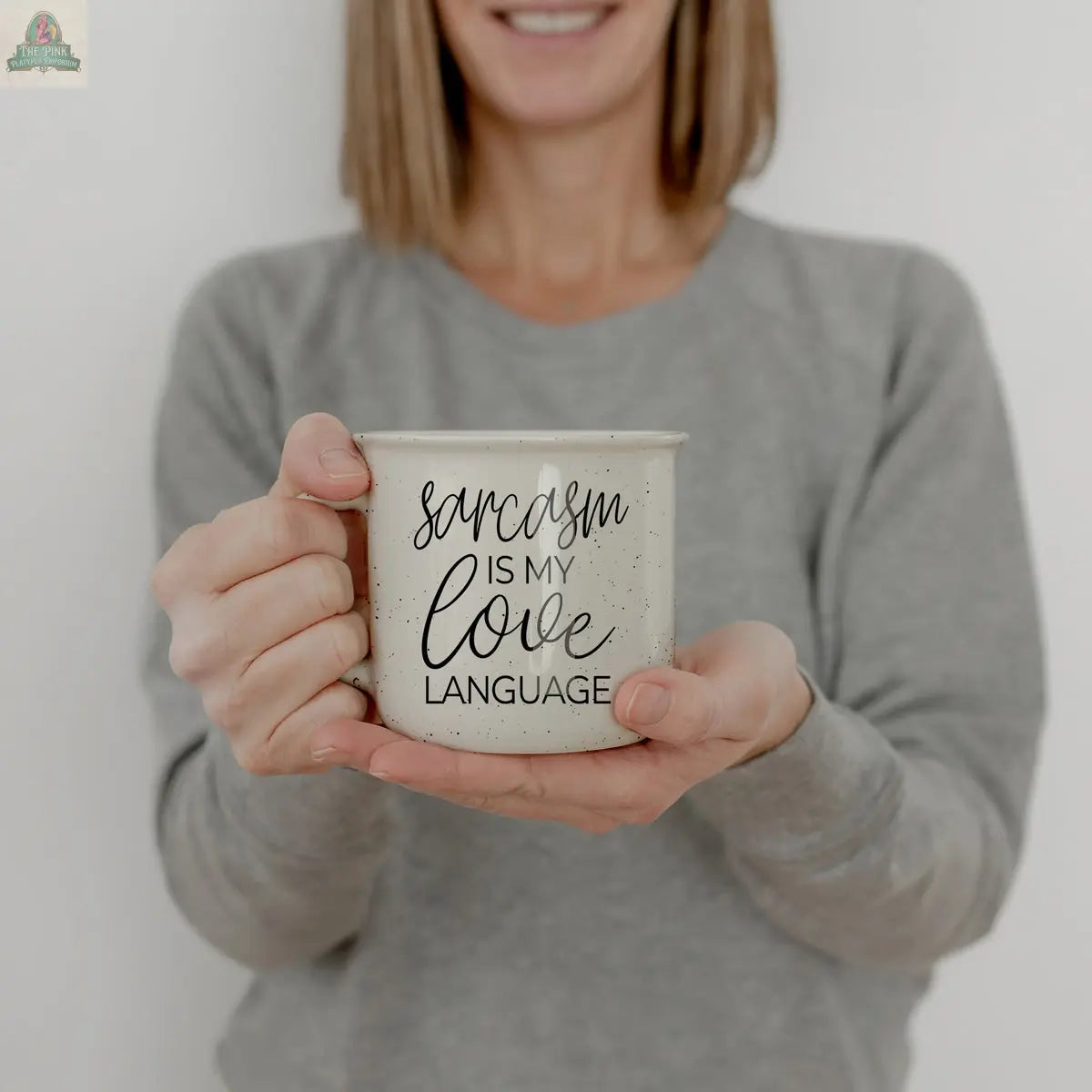 A woman in a gray sweater smiles while holding the Sarcasm Love 14.5oz ceramic coffee mug, featuring bold black text saying "Sarcasm is my love language," against a plain white background.