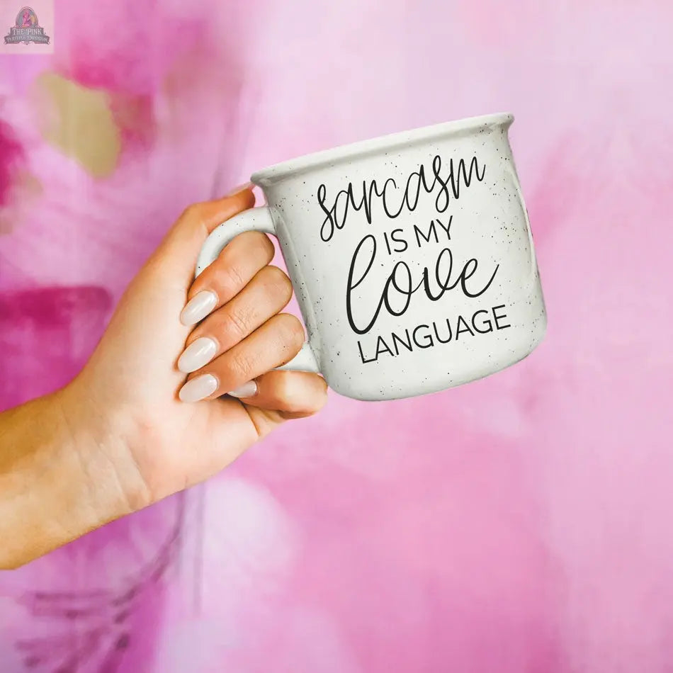 A hand with manicured nails holds the Sarcasm Love 14.5oz white ceramic mug against a pink background, featuring black text: "Sarcasm is my love language"—a perfect funny coffee mug for anyone with a witty personality.