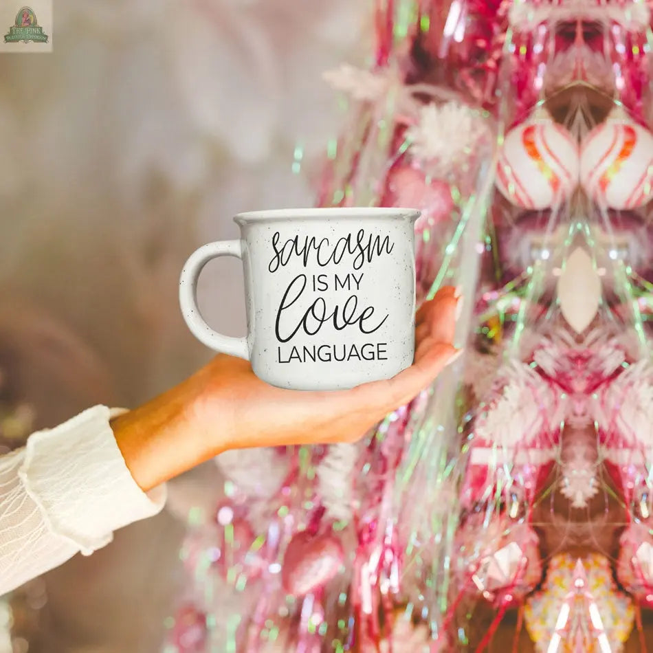 A hand holds a Sarcasm Love 14.5oz ceramic mug with "Sarcasm is my love language" text in front of a pink Christmas tree adorned with ornaments and tinsel.
