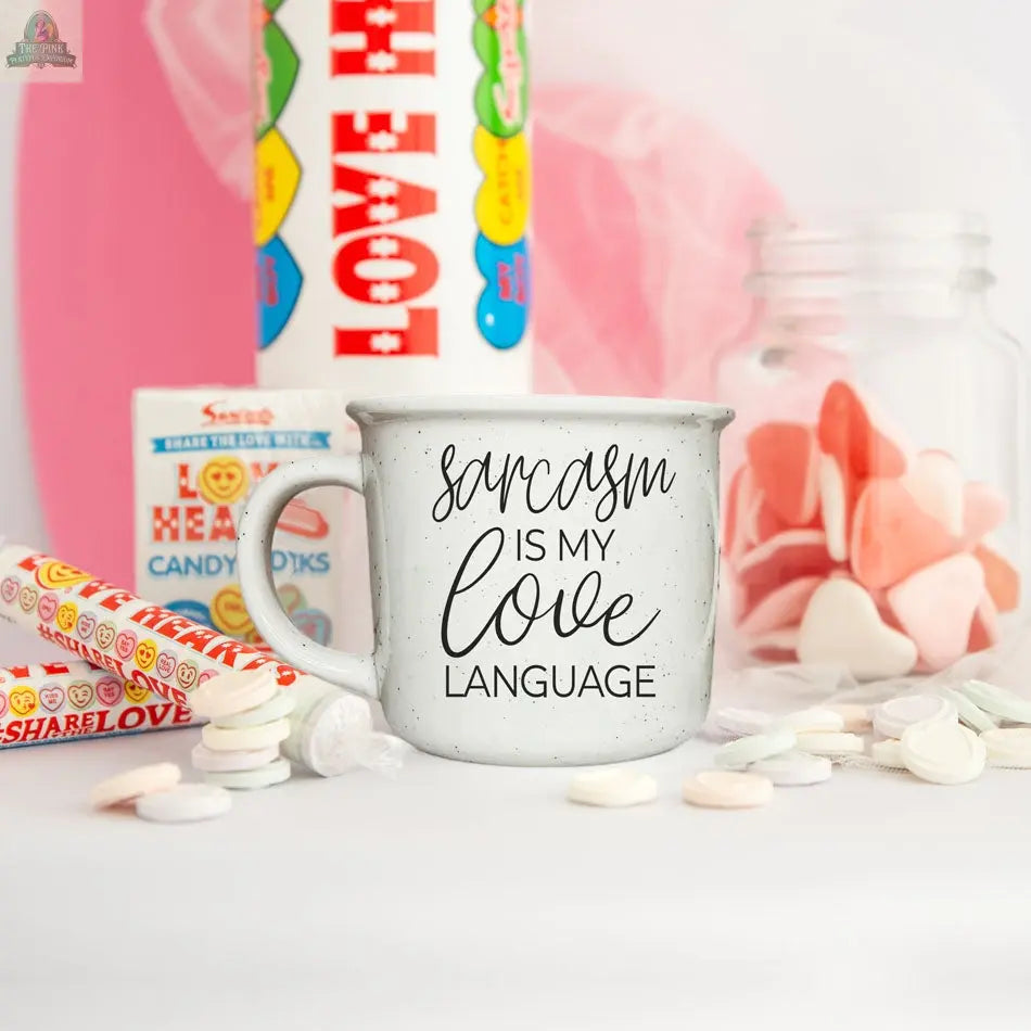 The Sarcasm Love 14.5oz mug, featuring “sarcasm is my love language,” sits on a white surface with candy hearts, Love Hearts rolls, a jar of heart-shaped candies, and a large red heart in the background.