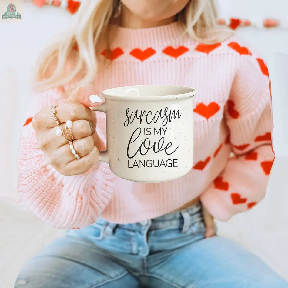 A woman in a pink sweater with red hearts playfully holds the Sarcasm Love 14.5oz ceramic mug, which reads “Sarcasm is my love language,” showing off her rings and blue jeans in this fun coffee mug moment.