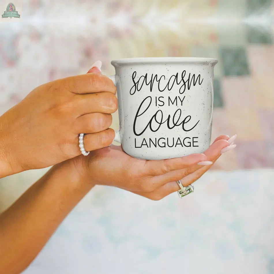 A person with manicured nails holds the Sarcasm Love 14.5oz ceramic mug featuring black text: "Sarcasm is my love language." This funny coffee mug pops against a softly blurred background.