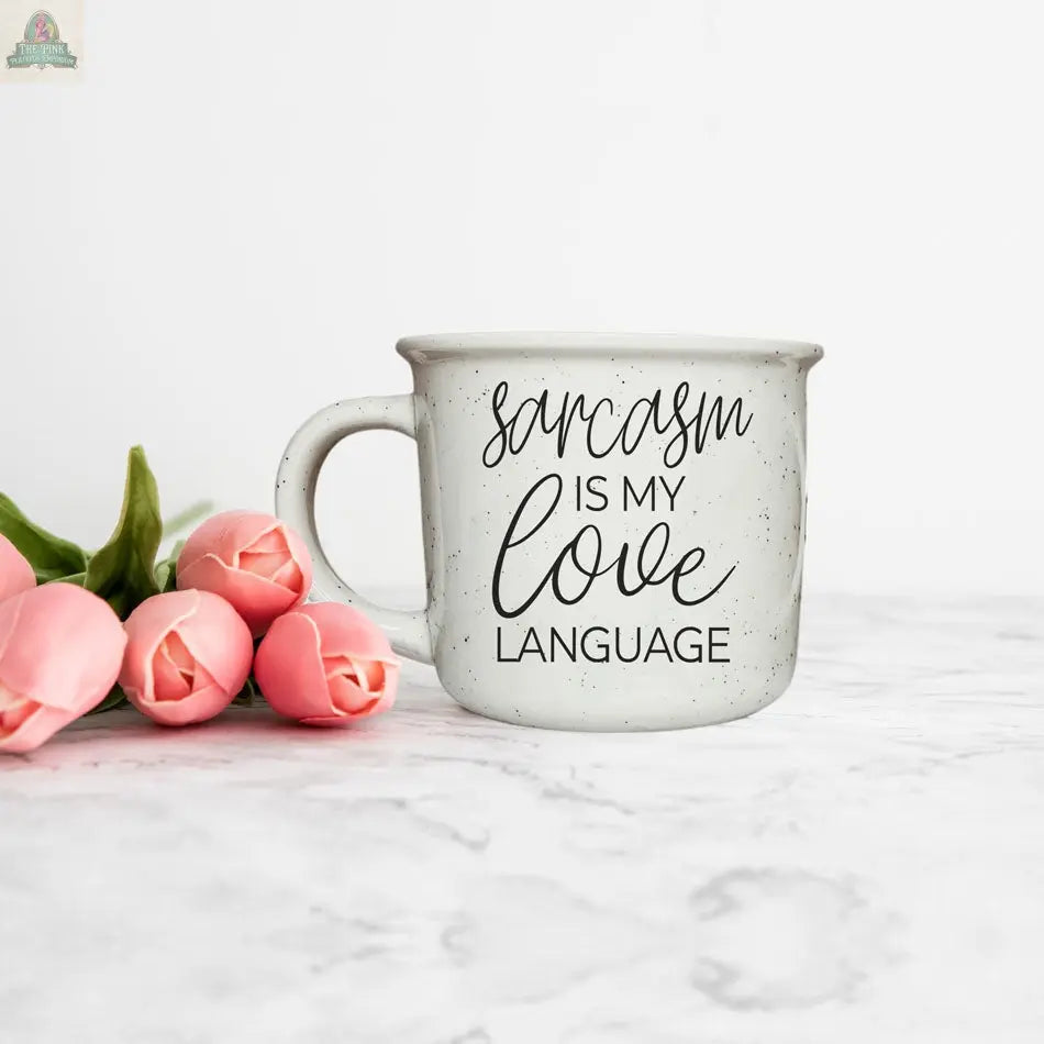 The Sarcasm Love 14.5oz white speckled ceramic mug, featuring the phrase “Sarcasm is my love language,” rests on a marble surface beside a small bunch of pink tulips.